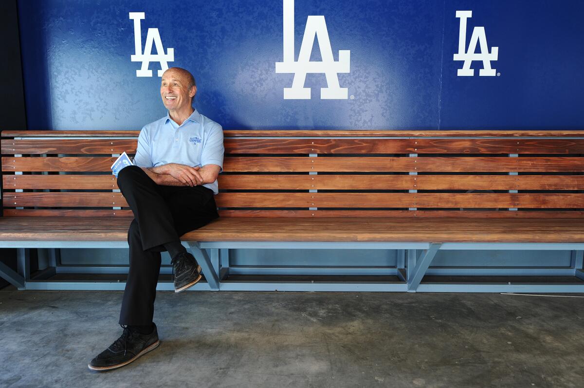 A man in a light blue shirt and black pants sits smiling on a wooden bench with a blue wall and the Dodgers logo behind him.