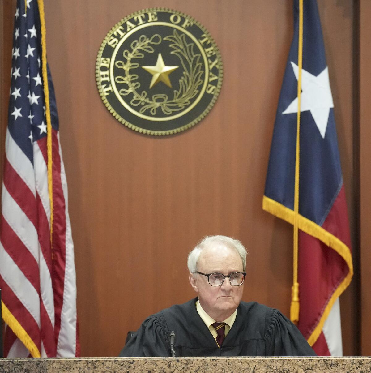 Judge David Peeples sits below a Texas state seal flanked by two flags.