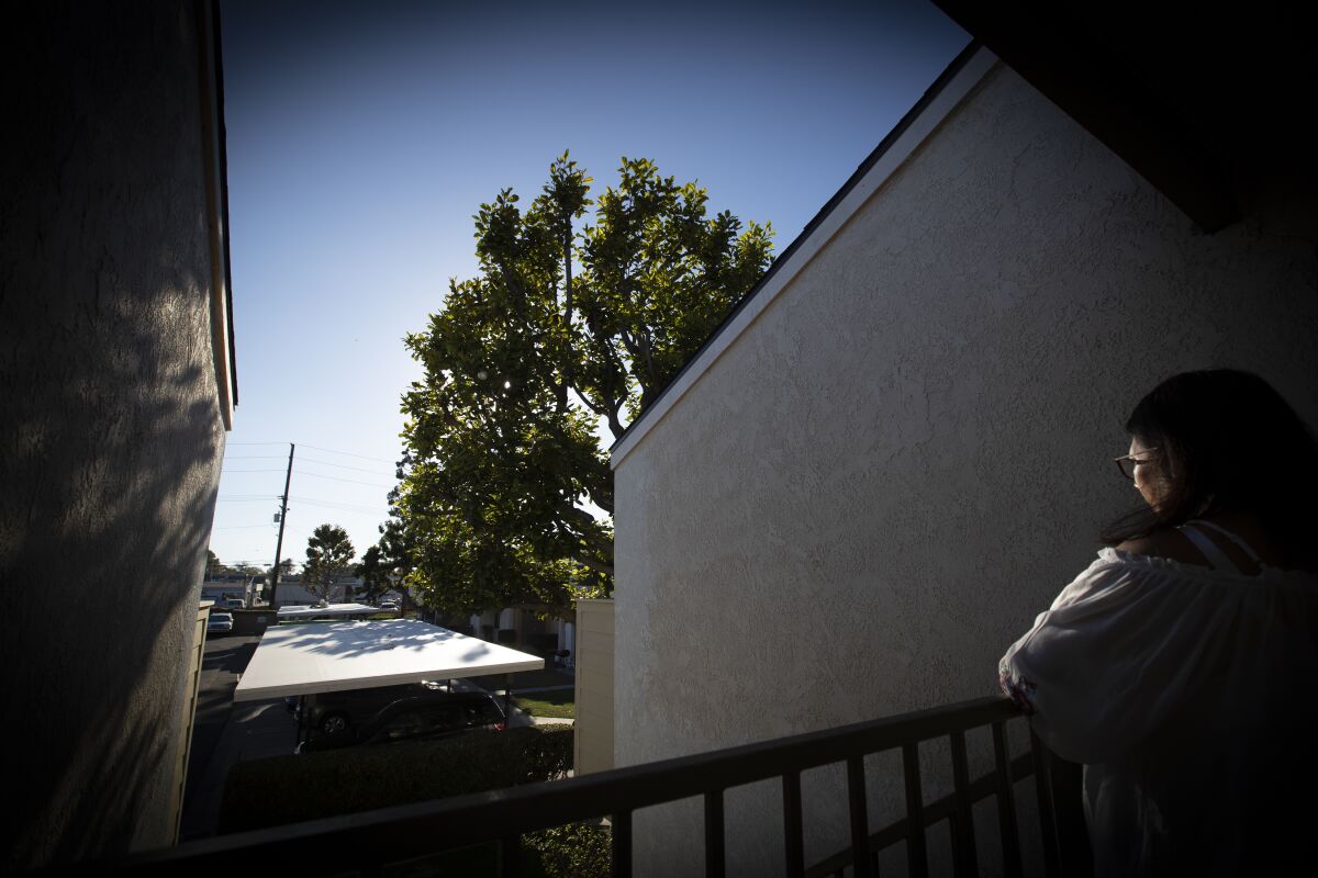 A woman stands looking out from a railed walkway of a stucco apartment building. 