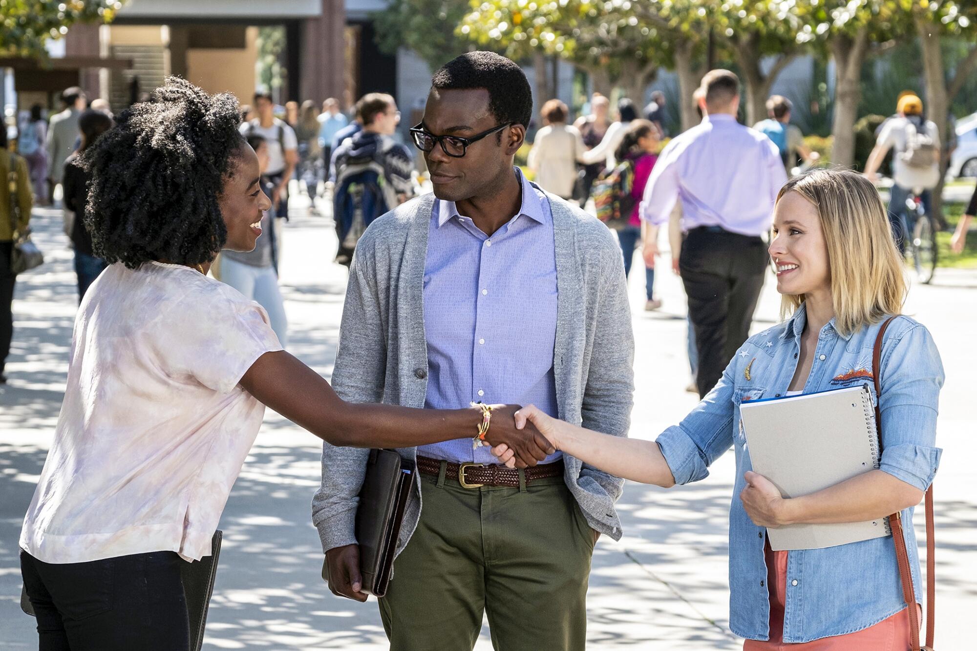 Two women shake hands as a man looks on.