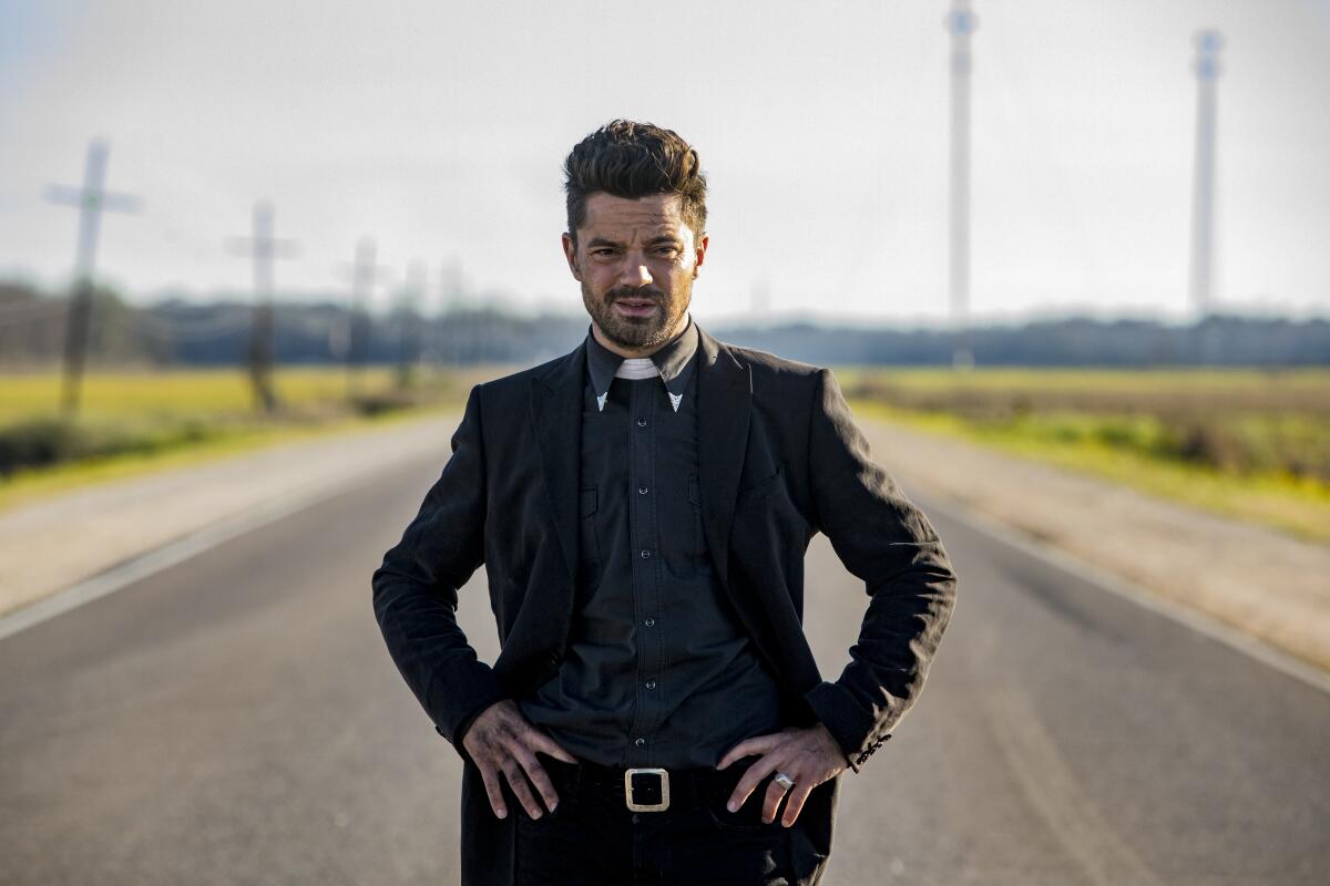 A priest wearing a collar stands with his hands on his hips in the middle of a rural road.