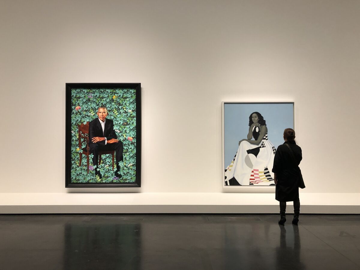 A woman wearing a black ensemble stands before large, colorful paintings of Barack and Michelle Obama