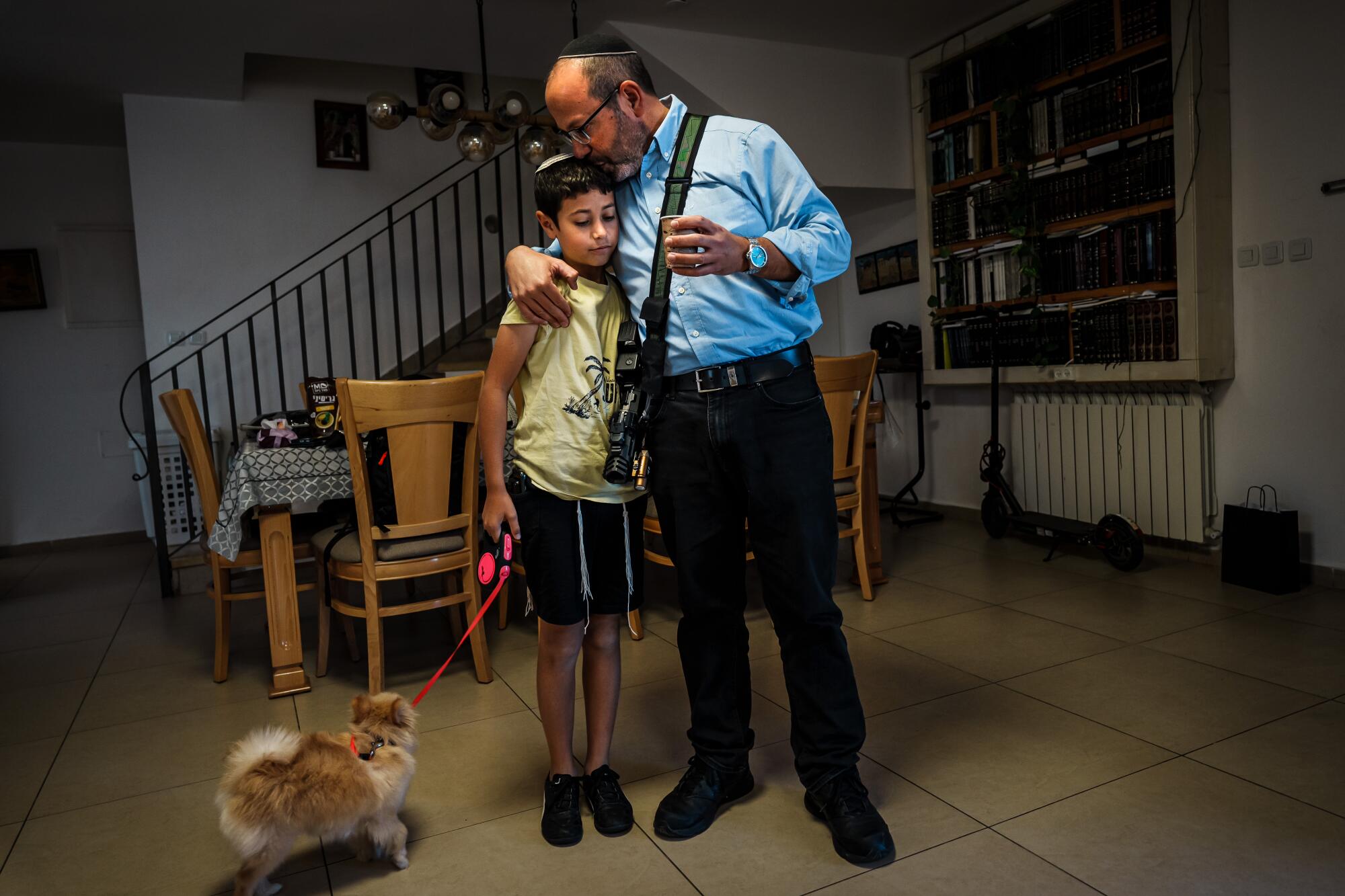 A man, wearing a long-sleeved blue shirt, dark pants and an assault weapon, hugs a boy holding the leash of a dog in a home