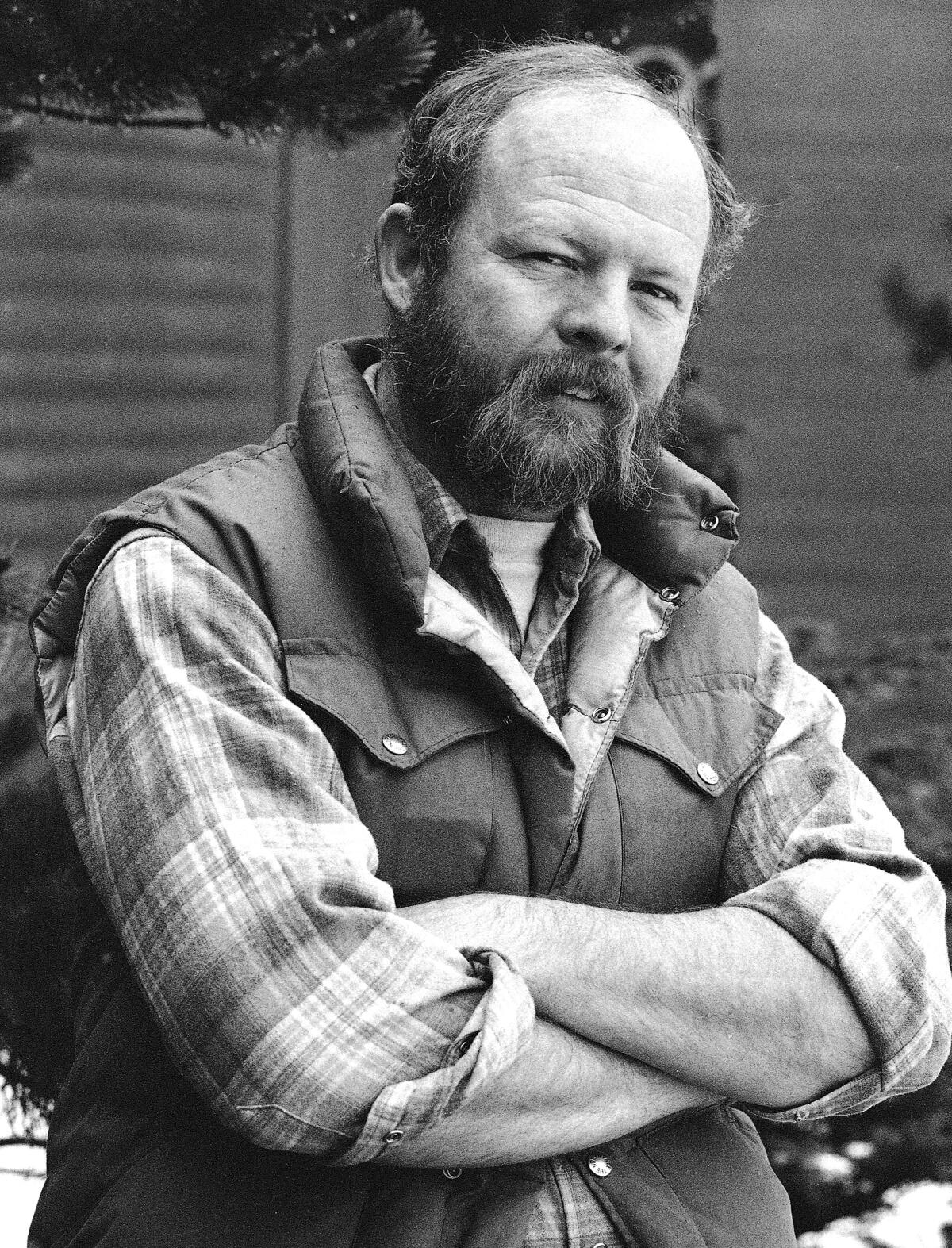 A man with a beard wearing a long-sleeved plaid shirt and a vest stands with his arms folded across his chest.