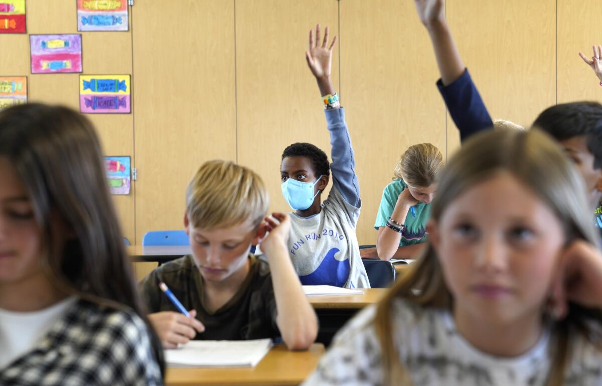 A masked boy raises his hand in class while unmasked students sit around him.
