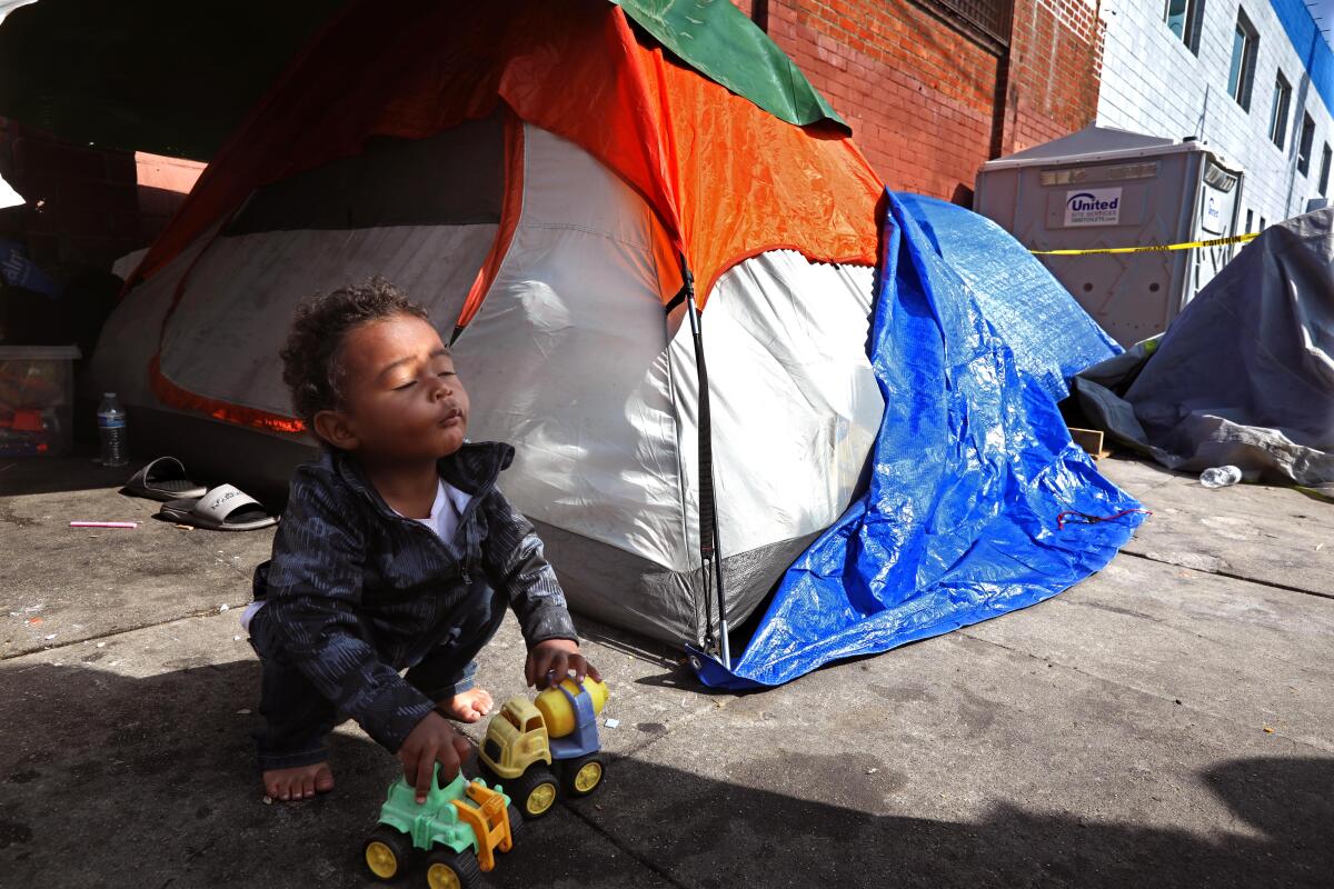 A young child with bare feet plays with small plastic toys on a sidewalk next to a tent.