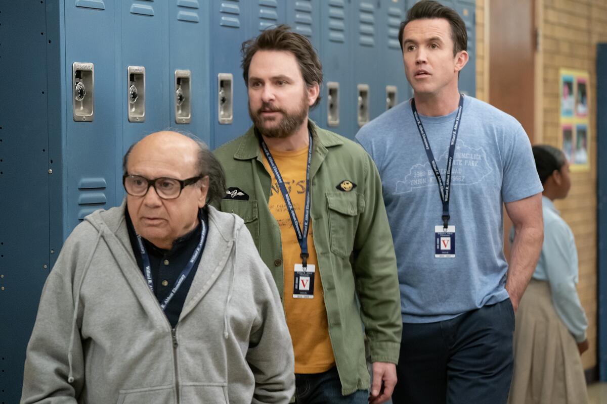 Three men lined up from shortest to tallest stand near a wall of blue school lockers.