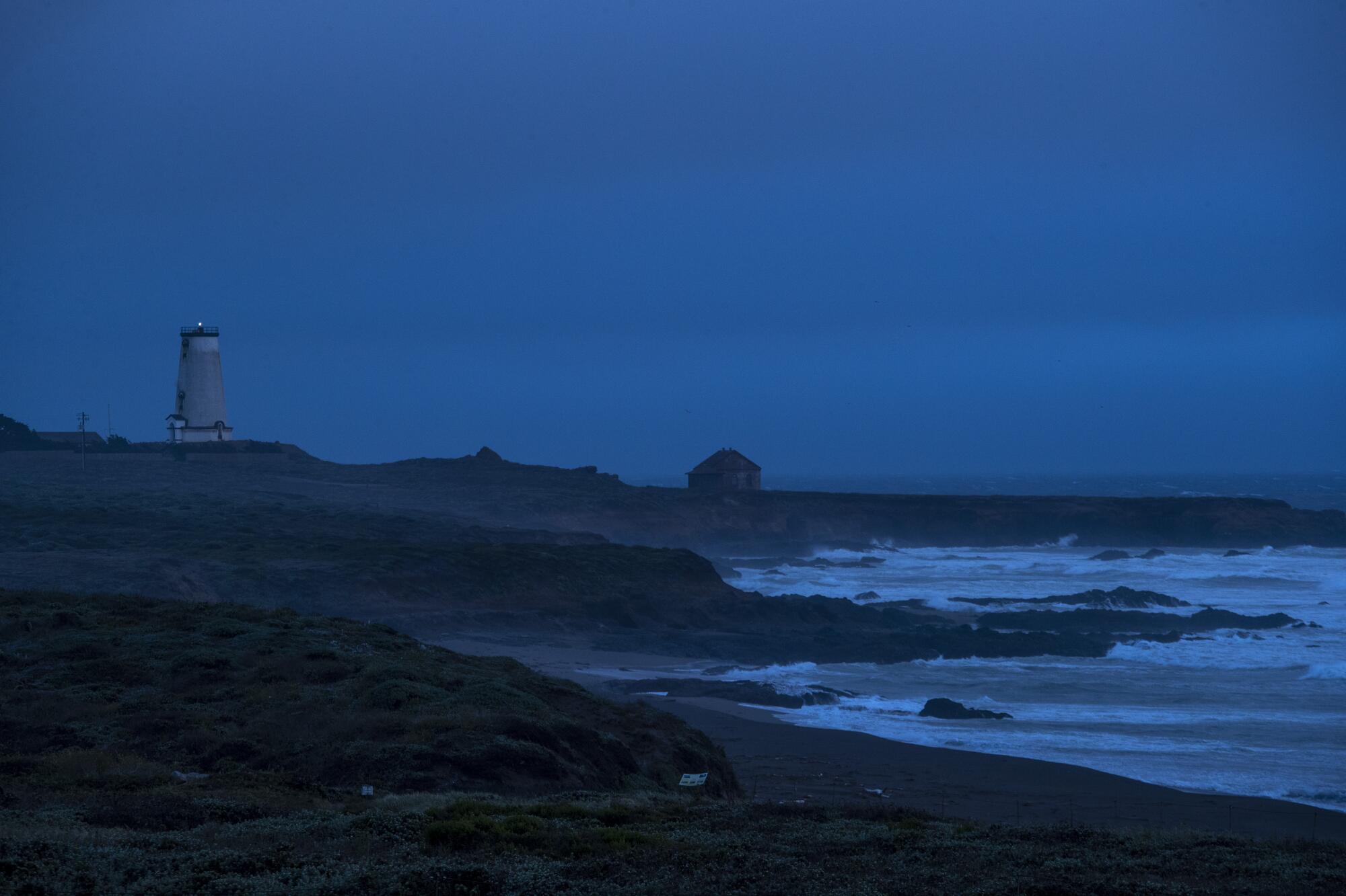 At dusk, a lighthouse and a lone building are silhouetted next to the surf.
