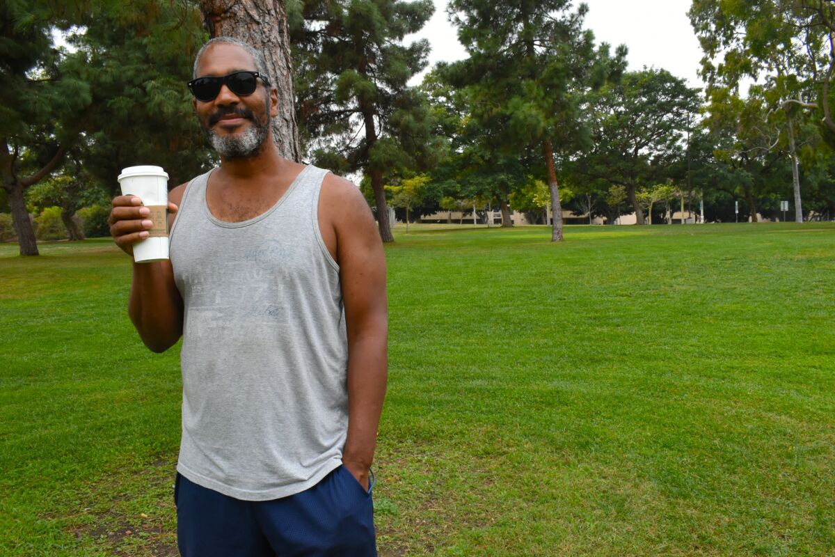 A man holding a hot drink and standing in the cut grass against a backdrop of evergreens