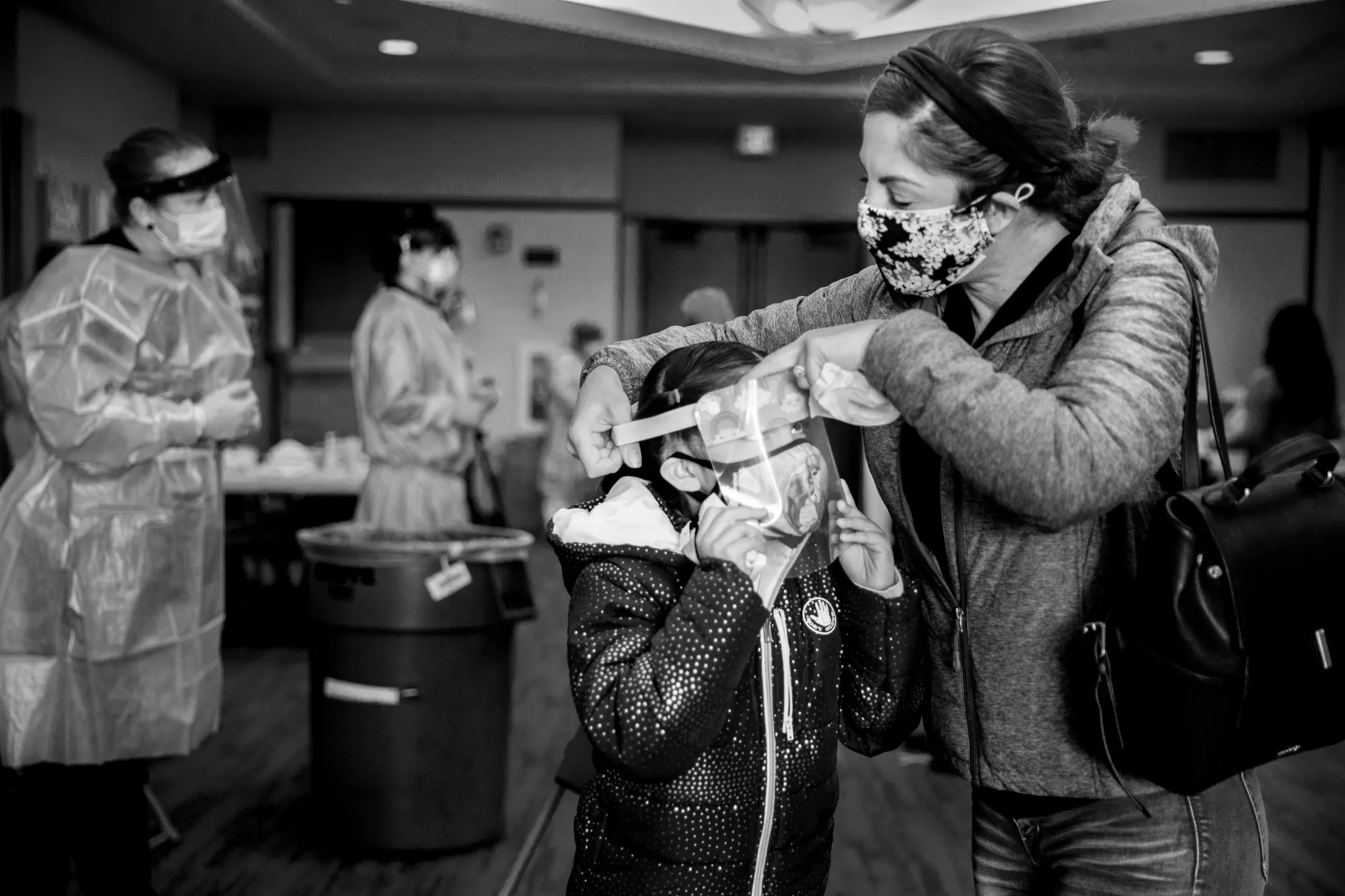 Black and white photo of mom helping daughter put on a Covid face shield