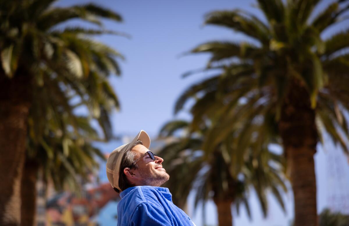 A man in sunglasses, cap and blue shirt is seen from the side, flanked by palm trees in the background