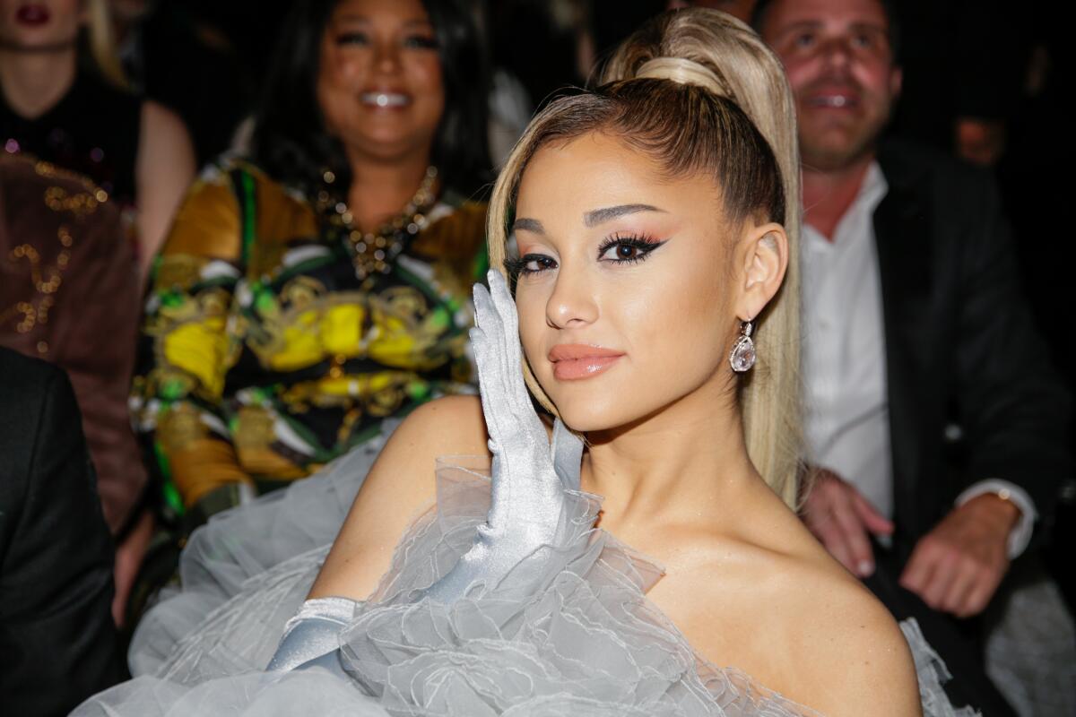 Ariana Grande wears a poofy gray gown and leans to her left while touching her cheek.