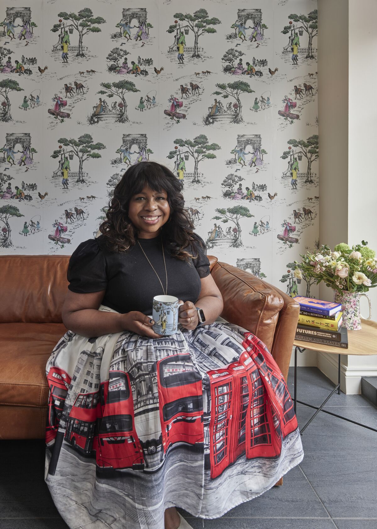 A woman sits on a couch in front of Harlem Toile wallpaper, in her home in London. 