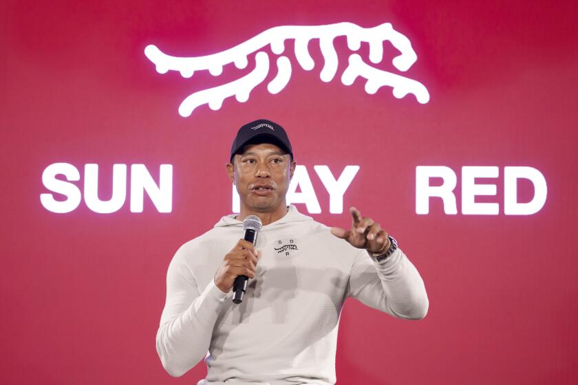Tiger Woods points and holds a microphone, standing in front of a red backdrop with the words 'Sun Day Red' and a tiger logo.