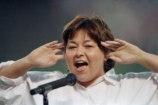 FILE - In this July 25, 1990, file photo, comedienne Roseanne Barr holds her fingers in her ears as she screams the National Anthem between games of the San Diego Padres and the Cincinnati Reds doubleheader in San Diego, Calif. A Delaware school district played a parody version of the national anthem before a volleyball match, surprising spectators with Barr’s screeching 1990s rendition. The News Journal of Wilmington reports the Seaford School District apologized Wednesday, Sept. 25, 2019, for the shock at its Tuesday game with Milford High. Superintendent David Perrington says pre-game proceedings will be improved. Officials didn’t say how Barr’s version, performed before a San Diego Padres game, came to be used. (AP Photo/Joan Fahrenthold, File)