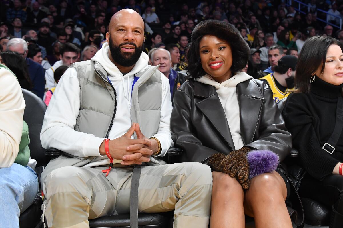 Common in a white hoodie, gray vest and gray pants sitting next to Jennifer Hudson in a hoodie and black jacket courtside