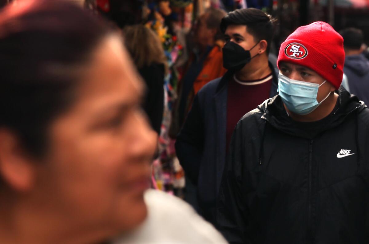 At left a woman stands blurred, close to camera, while two men in face masks walk down toward her on the right. 