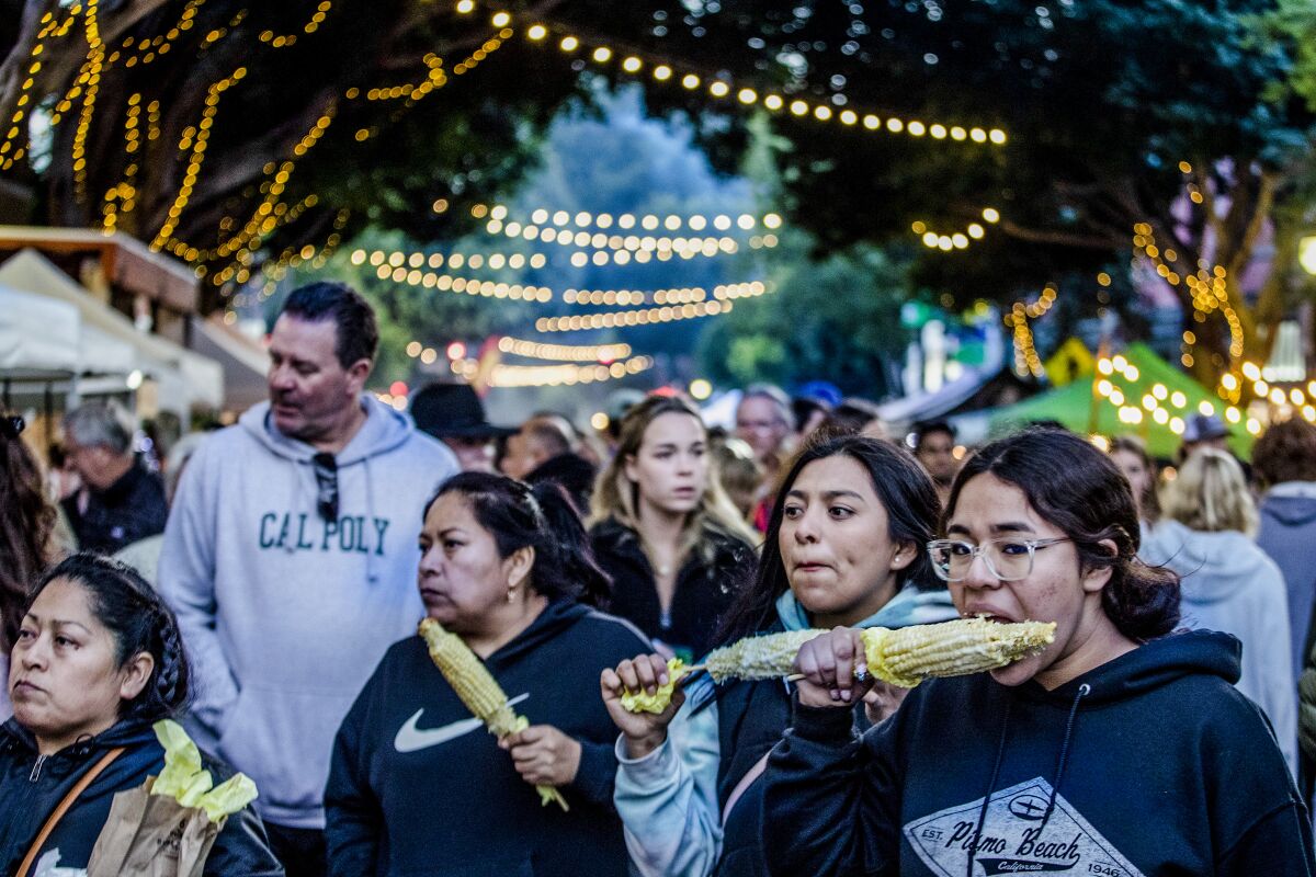 People eat corn on the cob outdoors under strings of lights at the weekly Downtown SLO Farmers' Market