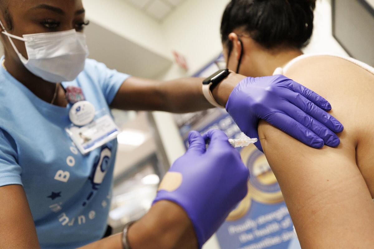 A woman in a blue shirt, wearing a mask, administers a shot on the upper arm of a patient