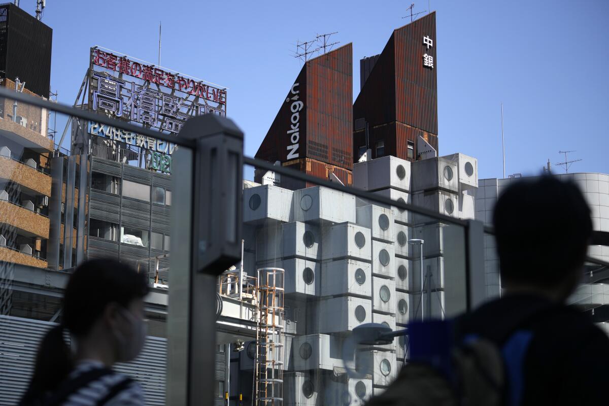 The Nakagin Capsule Tower's stacked-box architecture is seen through two pedestrians standing on the street.