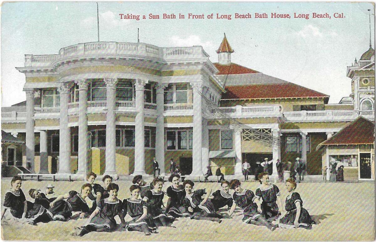 About 15 women sit or lie in the sand wearing black dresses and stockings in front of a neoclassical building