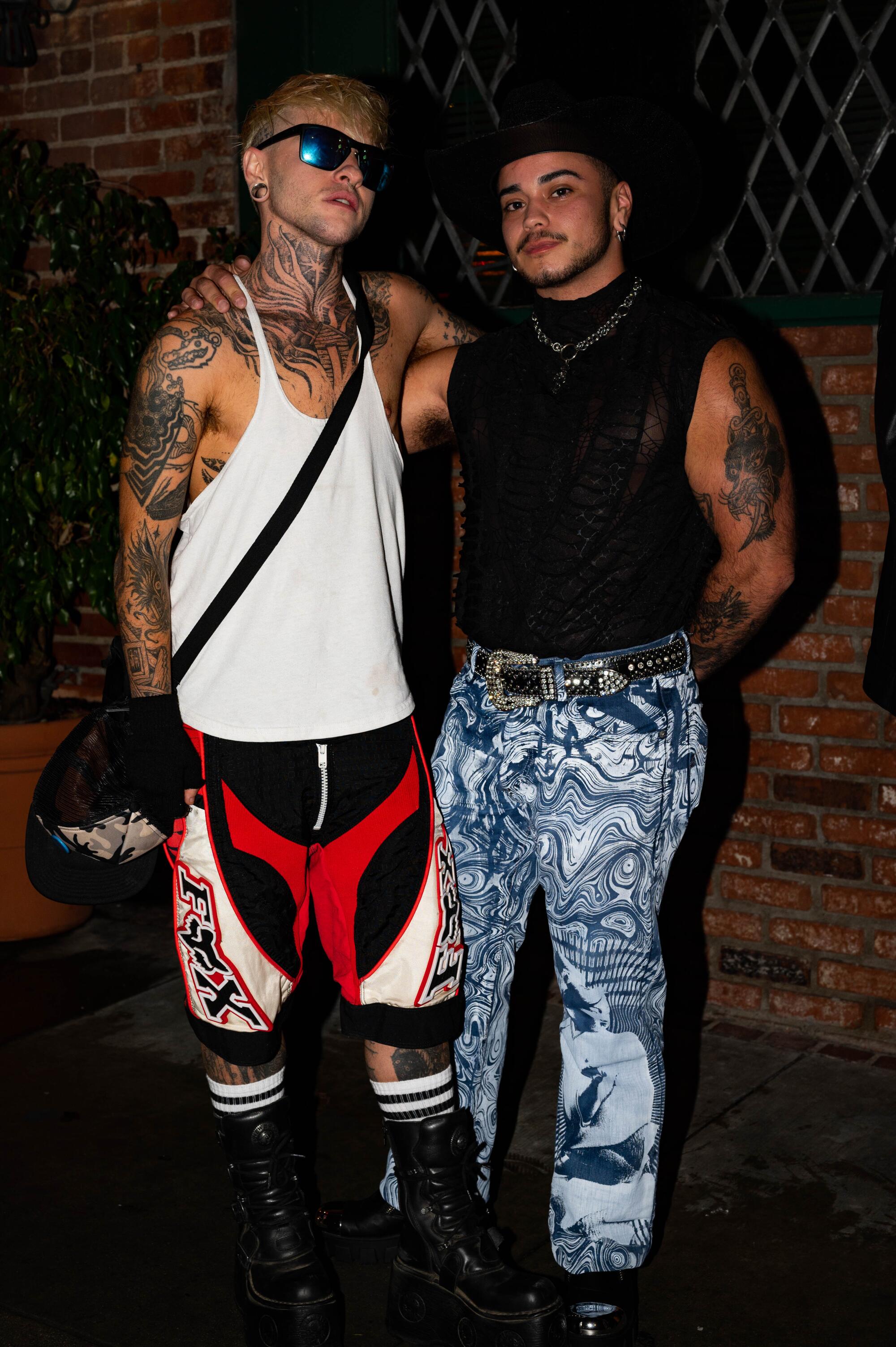 Two people wearing patterned pants pose side by side at night.