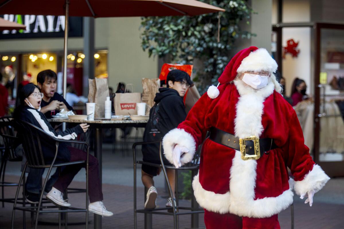 Santa Claus wearing a mask walks through an outdoor mall as a table of shoppers turn to look at him