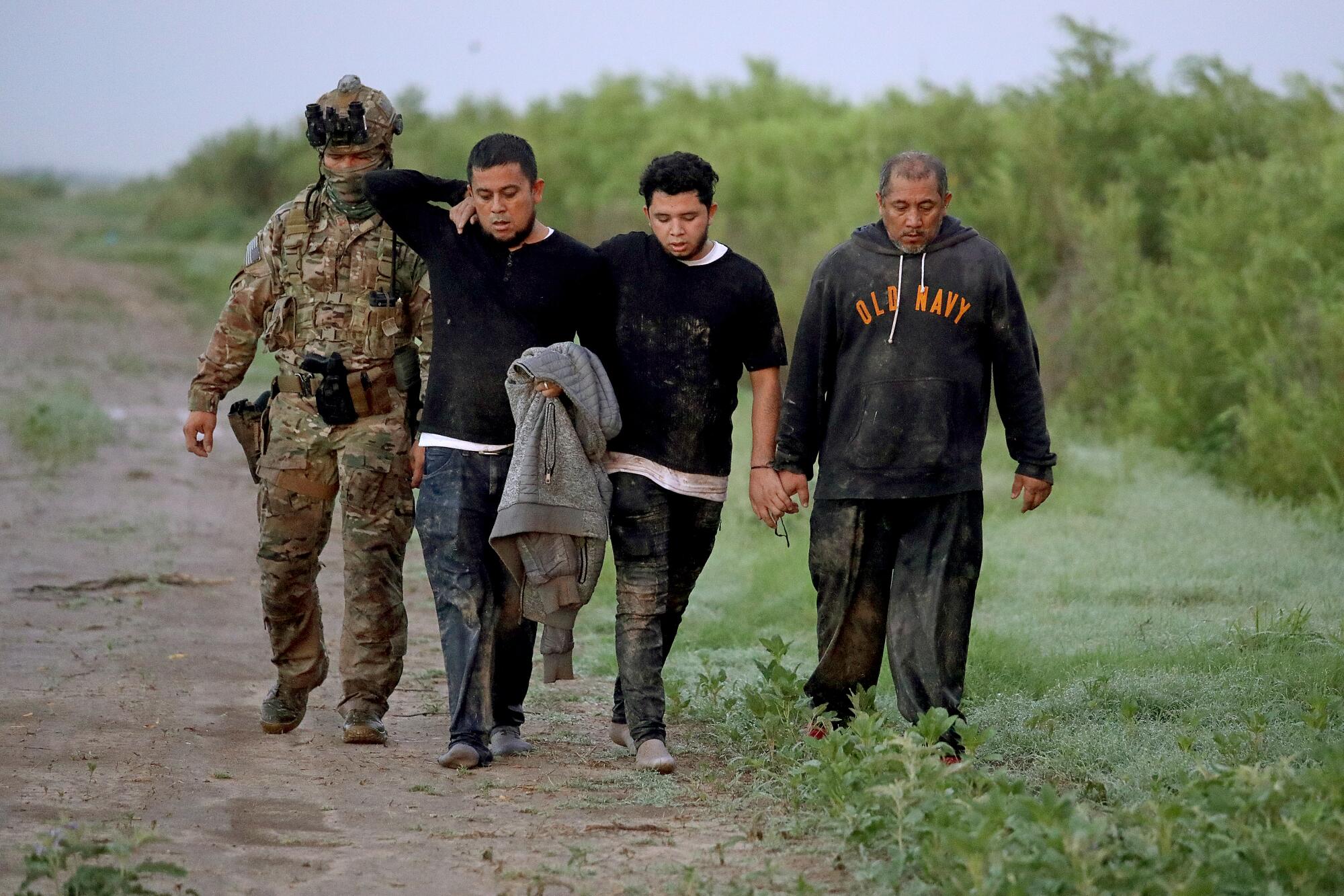A man in fatigues walks next to three other men in muddy clothes who are handcuffed