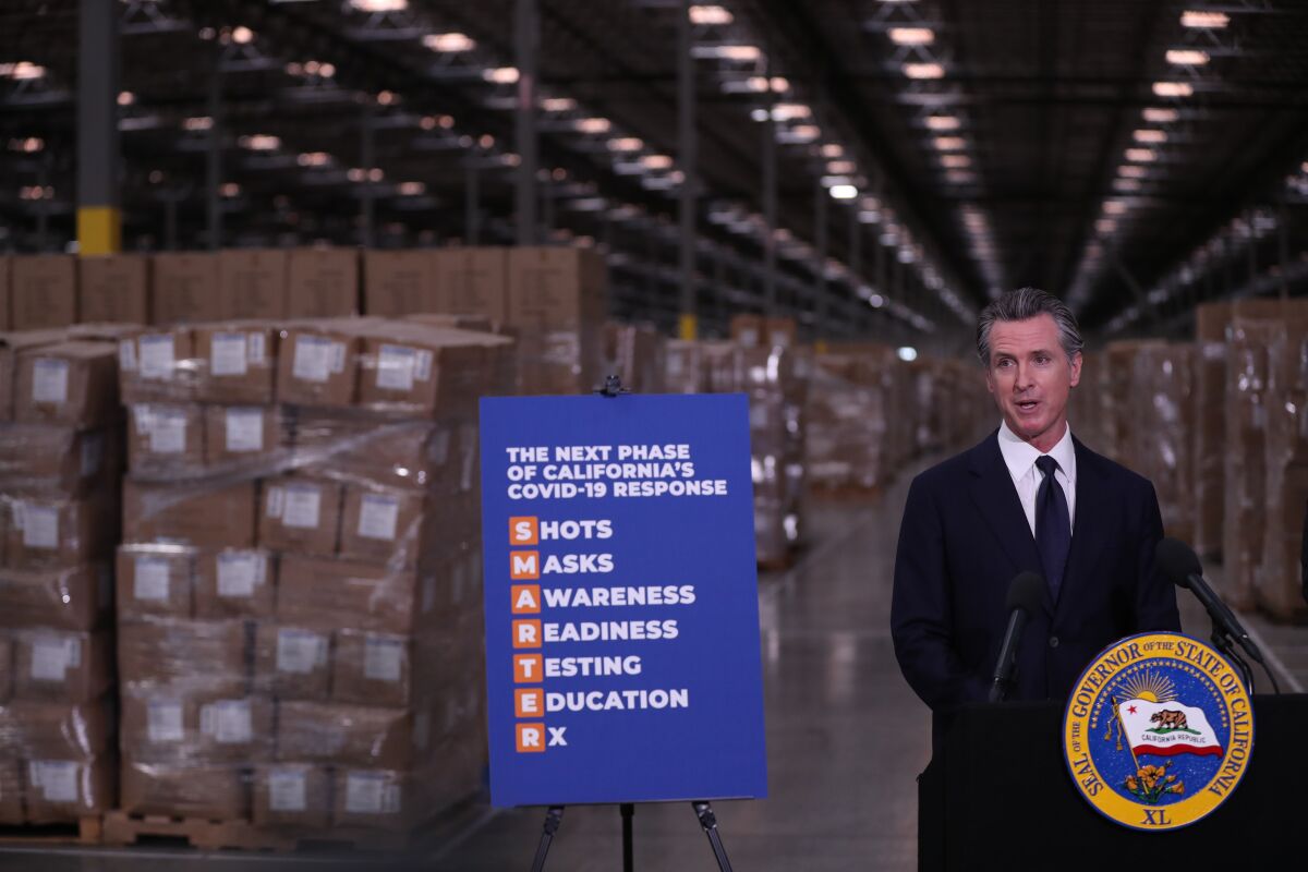 Governor Gavin Newsom speaks at a lectern in a warehouse with pallets of cardboard boxes behind him
