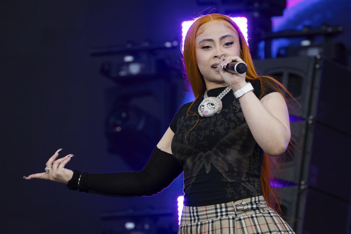 A woman in a black top and brown plaid skirt holds a microphone and extends her hand onstage