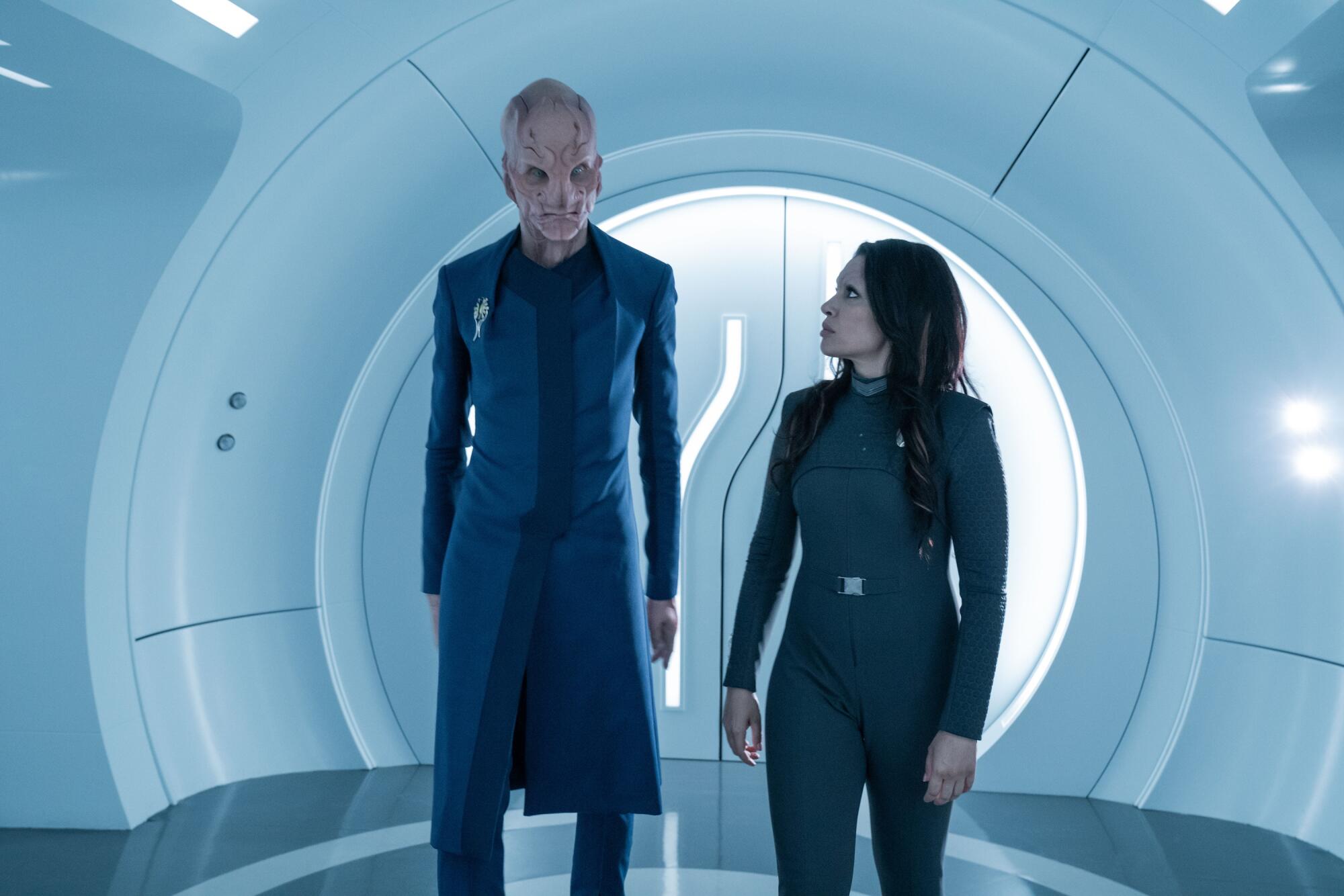 A tall, thin alien and a human woman walk through the tunnel of a spaceship.