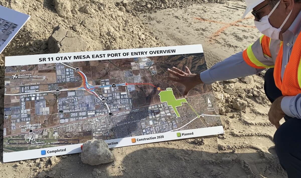 Mario Orso, in an orange vest, gestures to a map that is spread out on dirt with a rock holding it down.