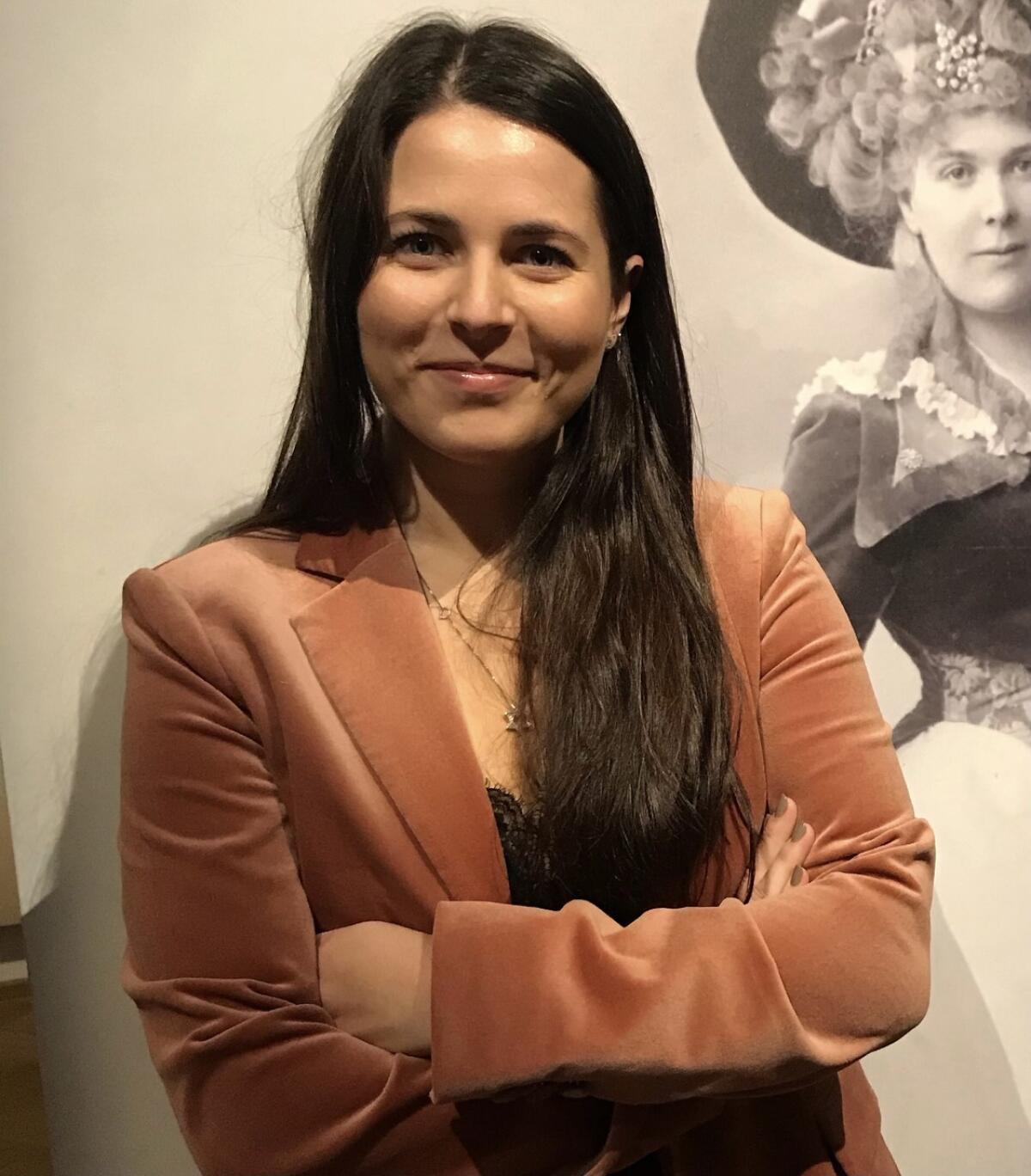 A woman in a peach velvet jacket crosses her arms and smiles.