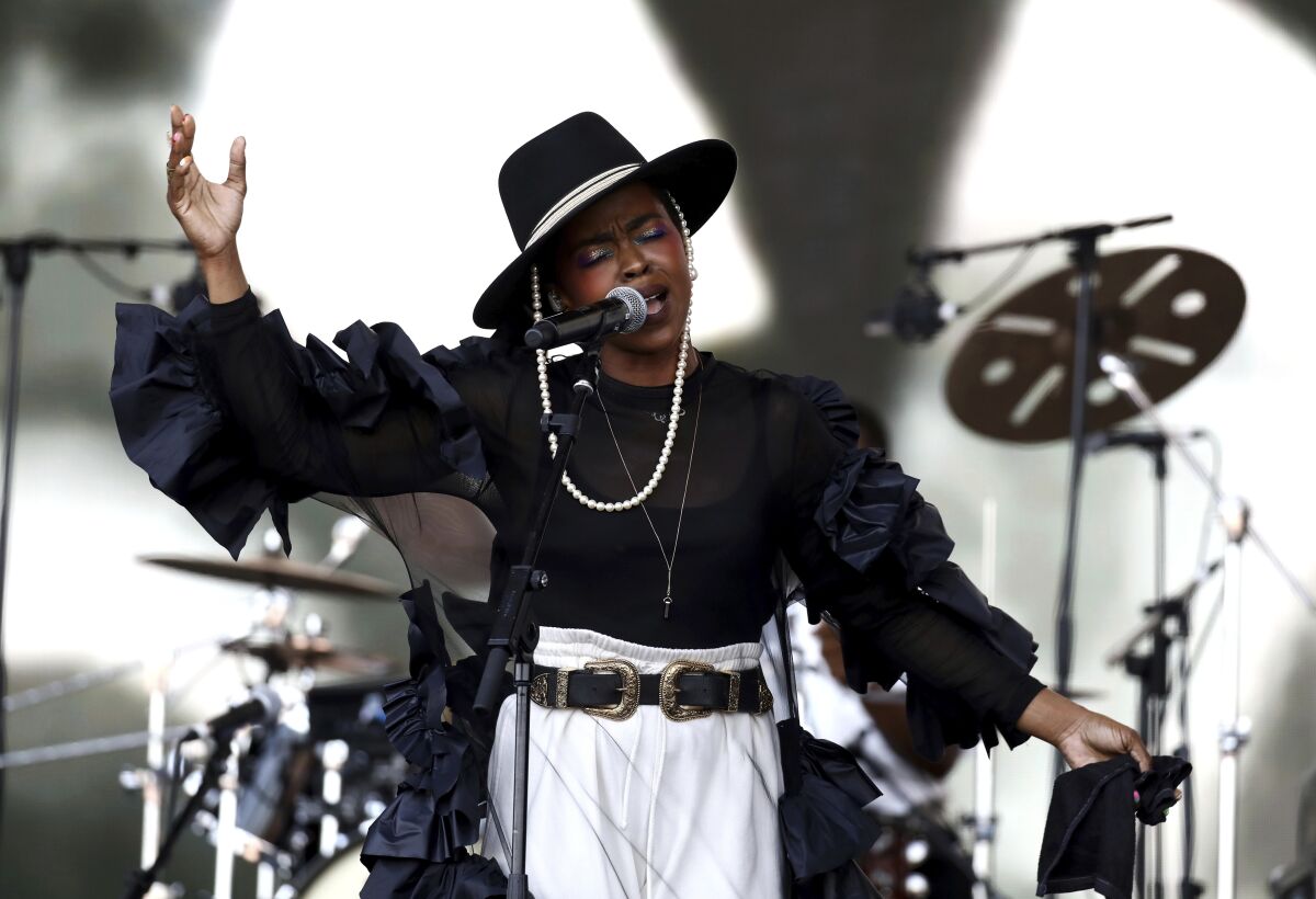 A woman in a black hat, black shirt and white pants singing in front of a microphone on a stage