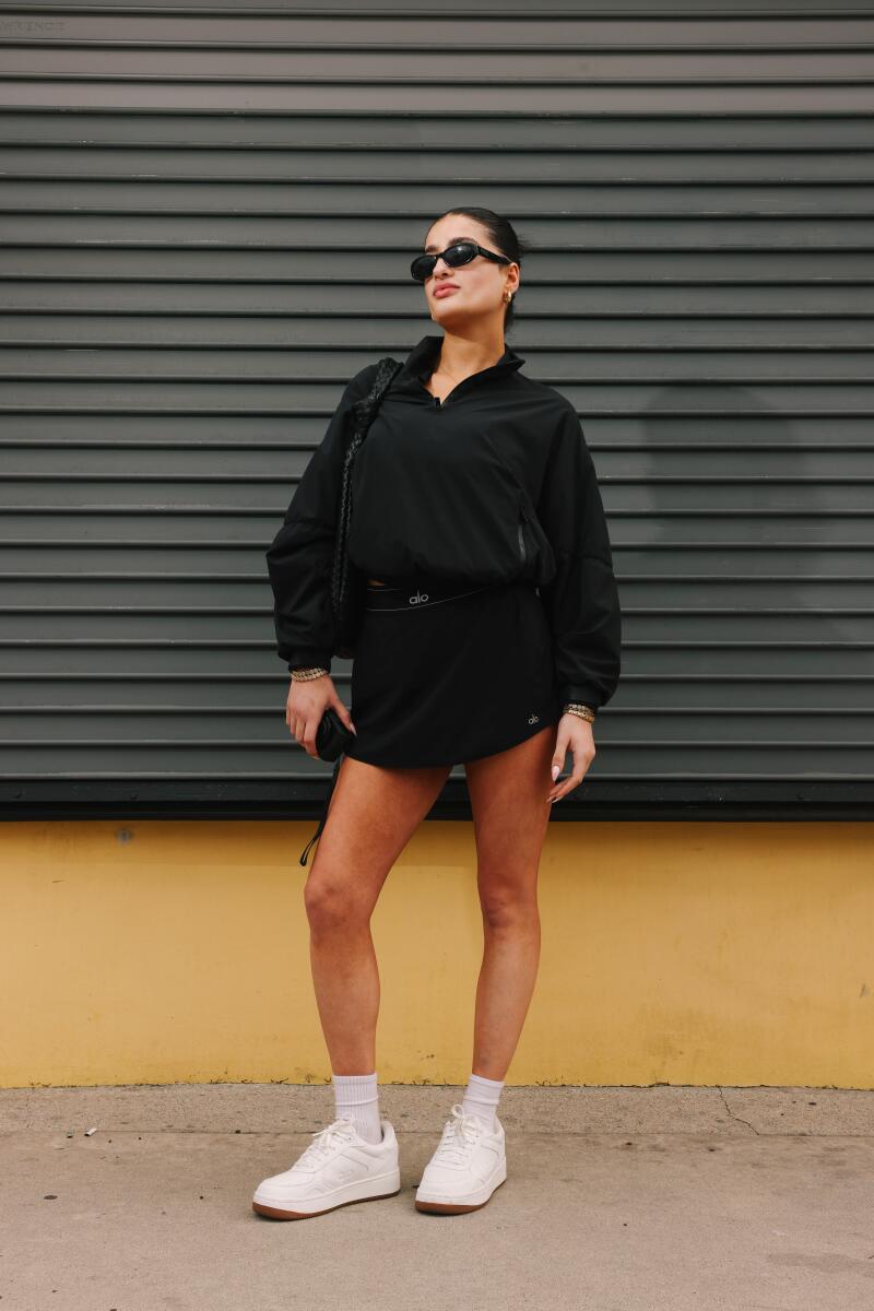A woman in black shorts and long-sleeved short, white sneakers and sunglasses stands outside a store
