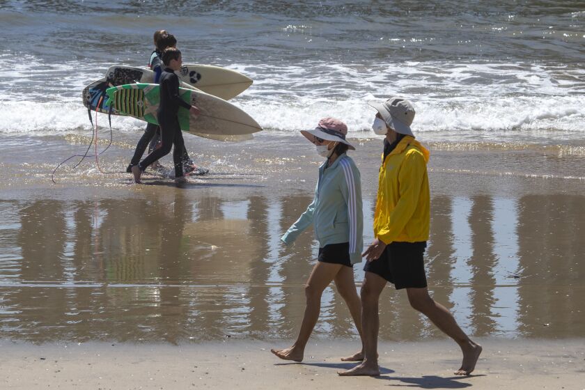 HUNTINTON BEACH, CA -- WEDNESDAY, APRIL 22, 2020: A trio of young surfers walks past a couple wearing protective masks as beach-goers enjoy warm summer-like weather amid state and city social distancing regulations mandated by Gov. Newsom in Huntinton Beach, CA, on April 22, 2020. (Allen J. Schaben / Los Angeles Times)