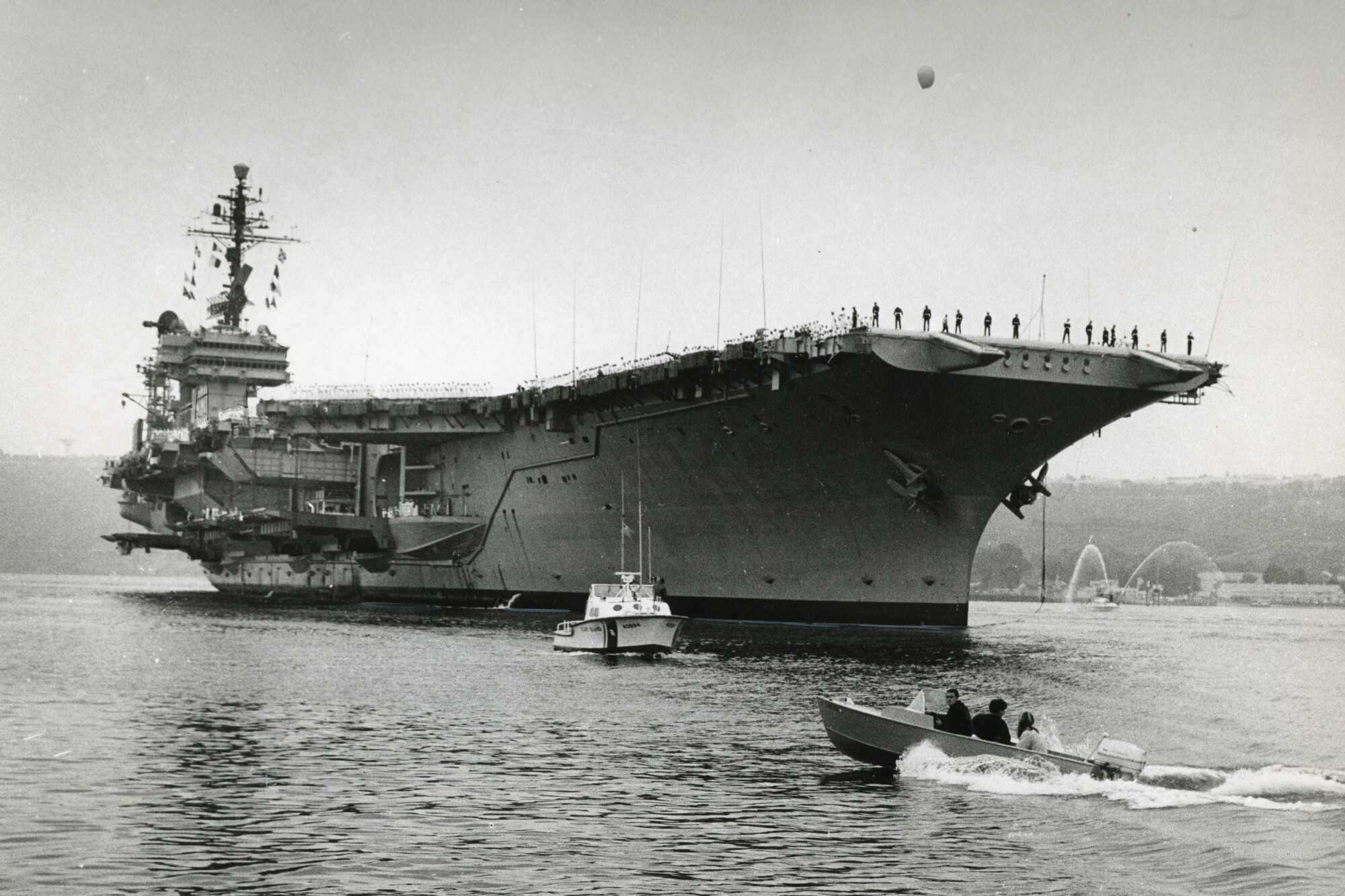 The aircraft carrier Kitty Hawk eases past Point Loma as an outboard motorboat roars beneath its bow in 1968.