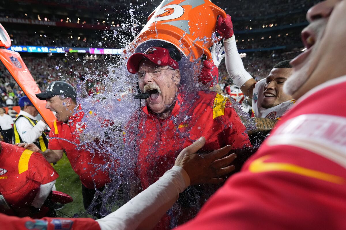 A man is doused in icy, purple liquid from a water cooler on the sideline in a football stadium.
