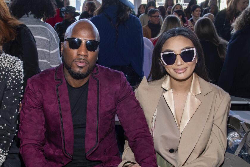 A bald Black man in a maroon suit and sunglasses sitting with an Asian woman with long hair in a beige suit and sunglasses