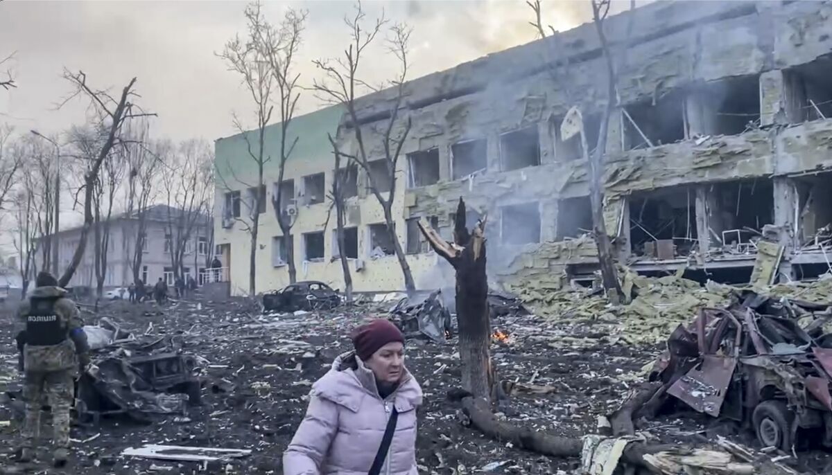 A person walks by bombed buildings and burned trees.