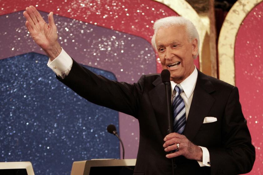 Bob Barker in a black suit and striped tie holding a microphone in his right hand and holding out his left arm