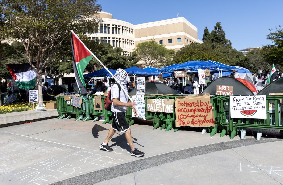 A person holding a Palestinian flag and wearing a kaffiyeh walks by an encampment at UC Irvine