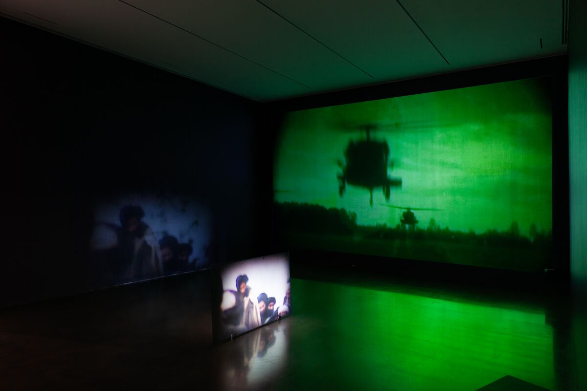 A dim gallery features a projection of a military helicopter bathed in green light and a monitor showing male figures