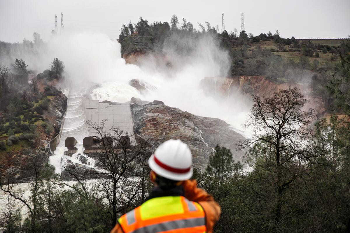 A man in an orange vest and hard hat looks toward a massive breach in a spillway of a dam.