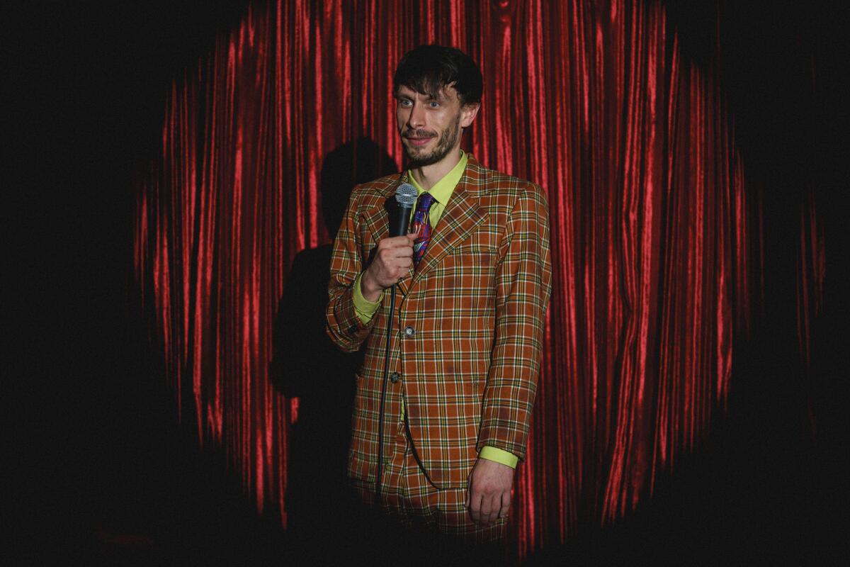 A man in a plaid suit holding a microphone onstage with a red curtain behind him.