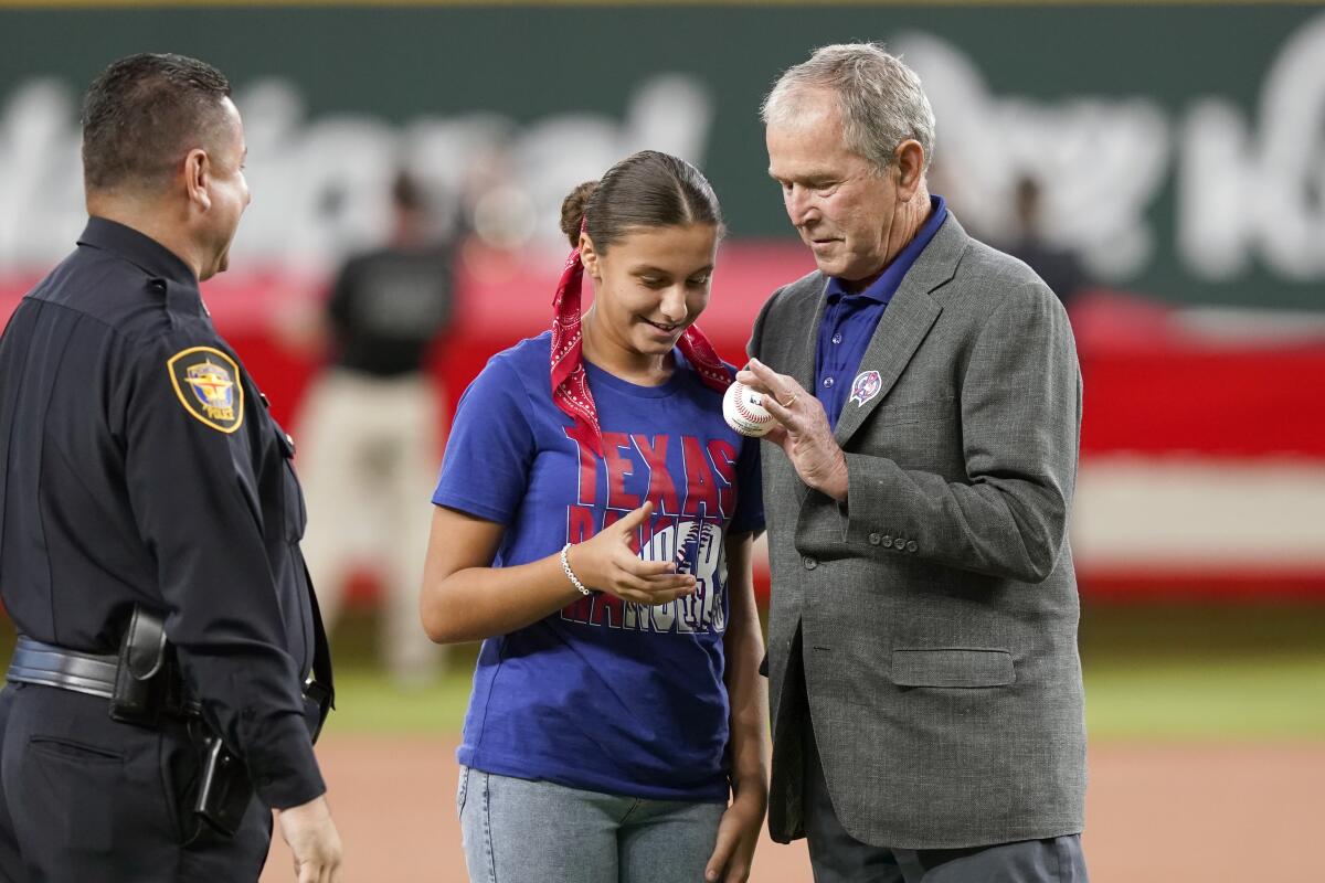 Former President George W. Bush, right, hands the ball to Andita Pollozani, center, for the ceremonial first pitch as her father, left, Fort Worth, Texas police officer Jimmy Pollozani, looks on to recognize the 21st anniversary of Patriot Day before a baseball game between the Toronto Blue Jays and the Texas Rangers in Arlington, Texas, Sunday, Sept. 11, 2022. (AP Photo/LM Otero)