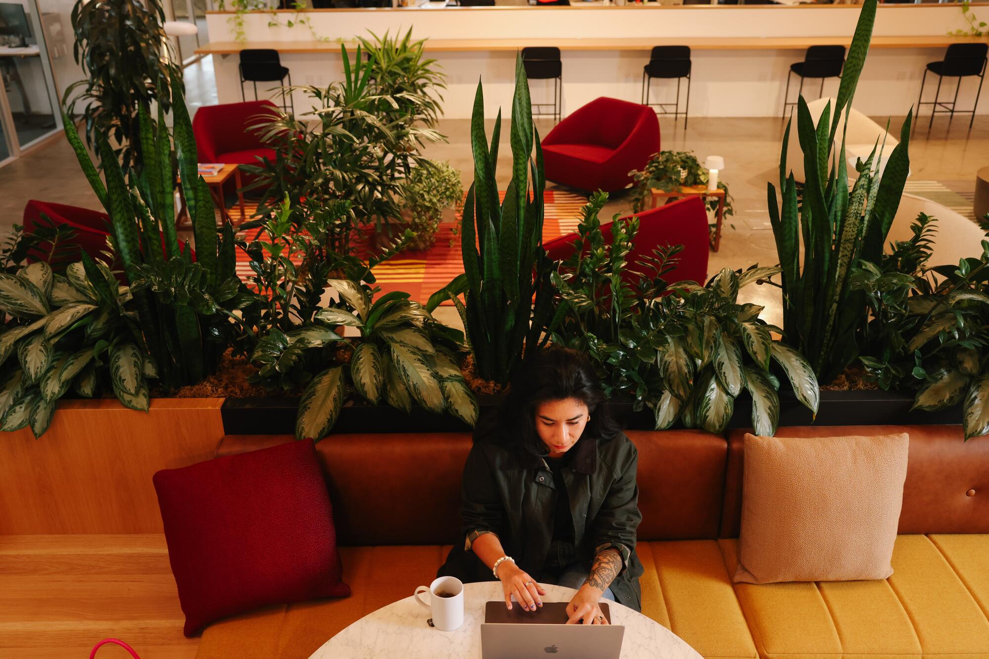 A lone woman seated on a sofa works at her laptop on a round table in an office setting.