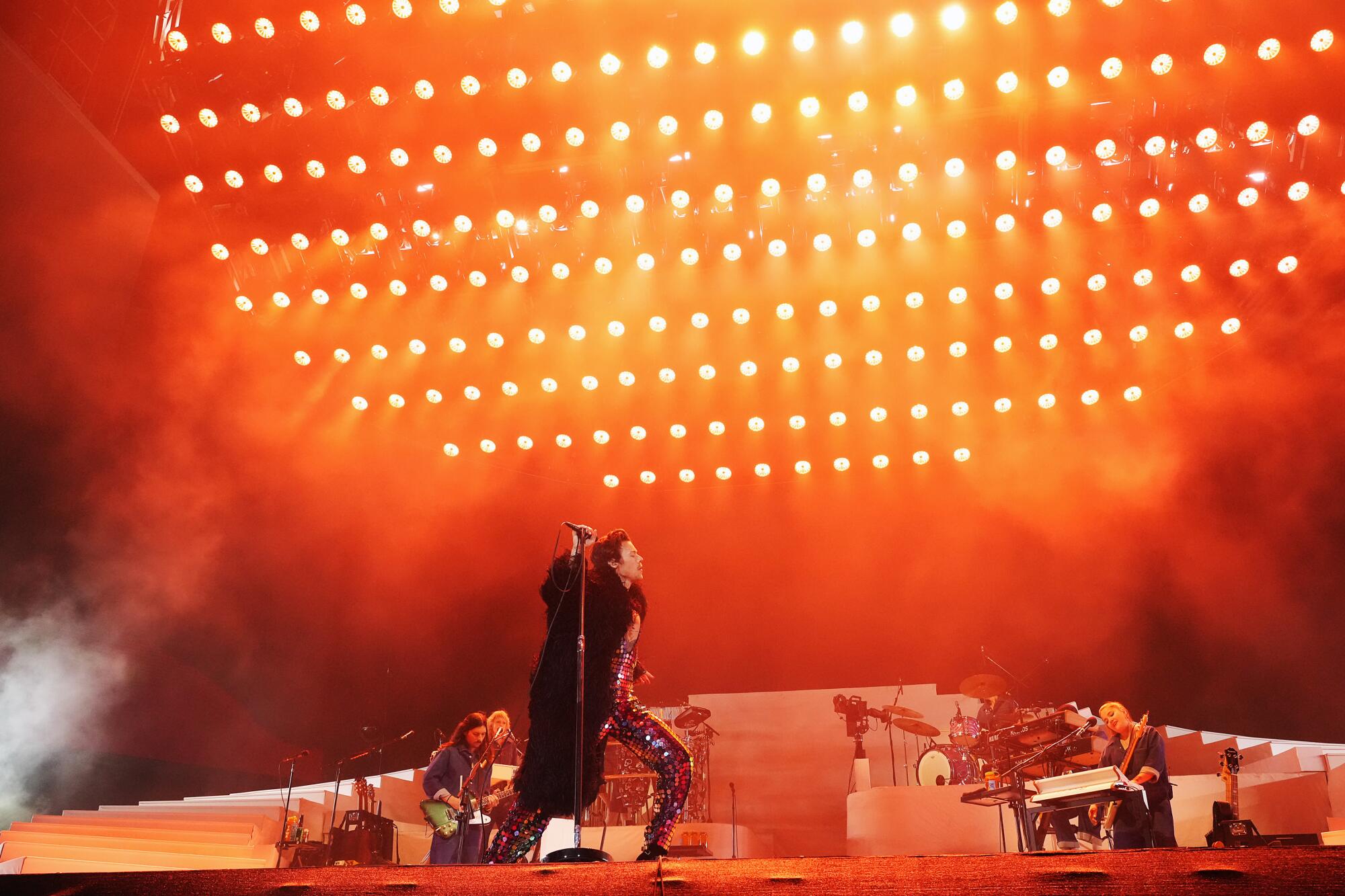 Harry Styles walks onstage in a coat and sparkly jumpsuit while rows of lights shine through smoke above him.