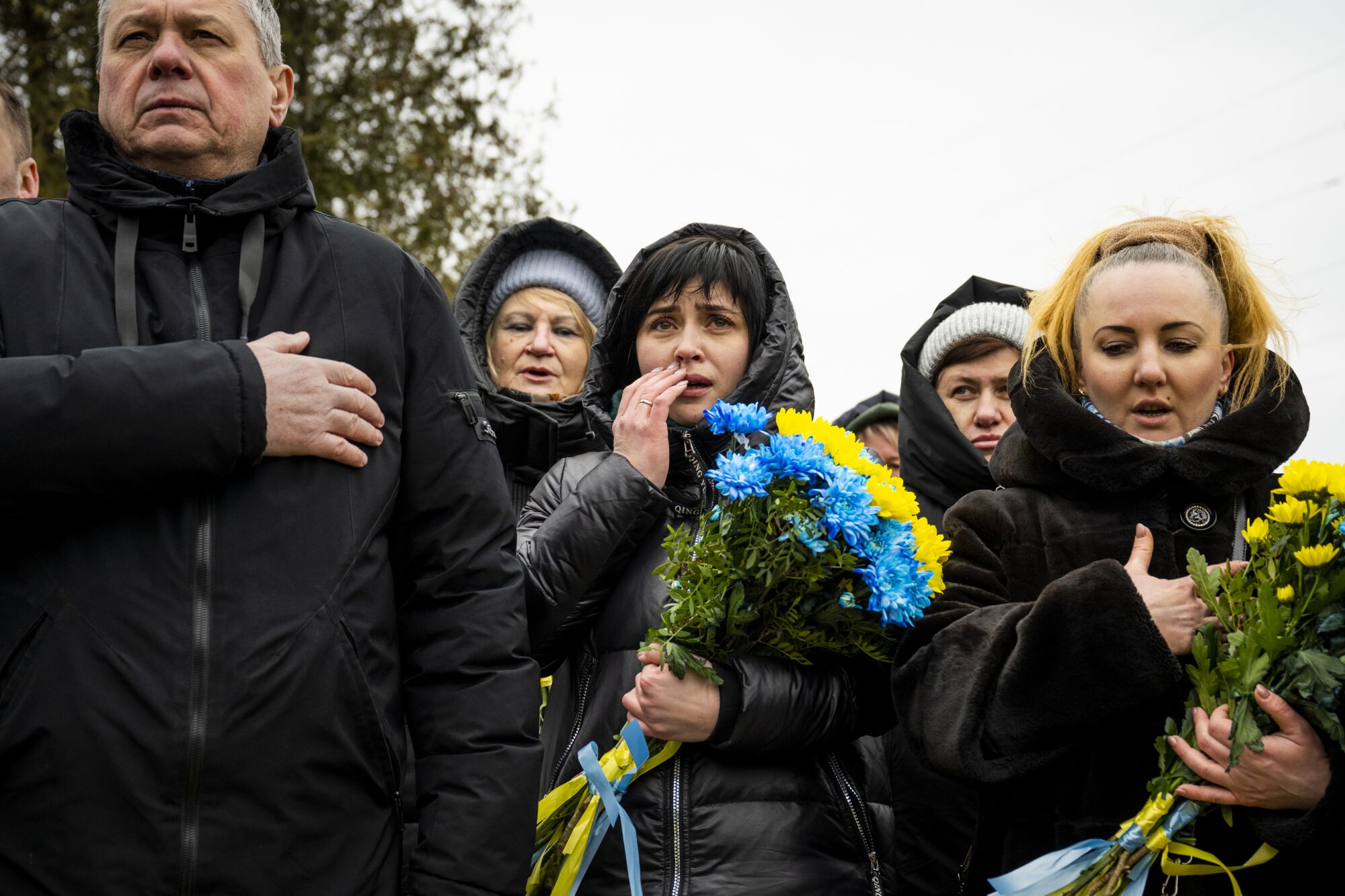 Five people, all in black winter clothes, stand together.  Some hold their hands over their hearts and some hold flowers. 