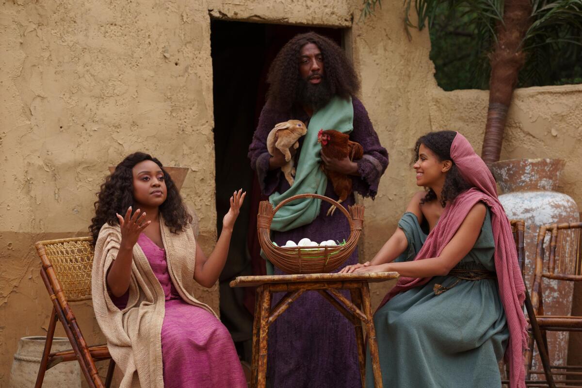 People in biblical clothing converse outside an earthen building. 